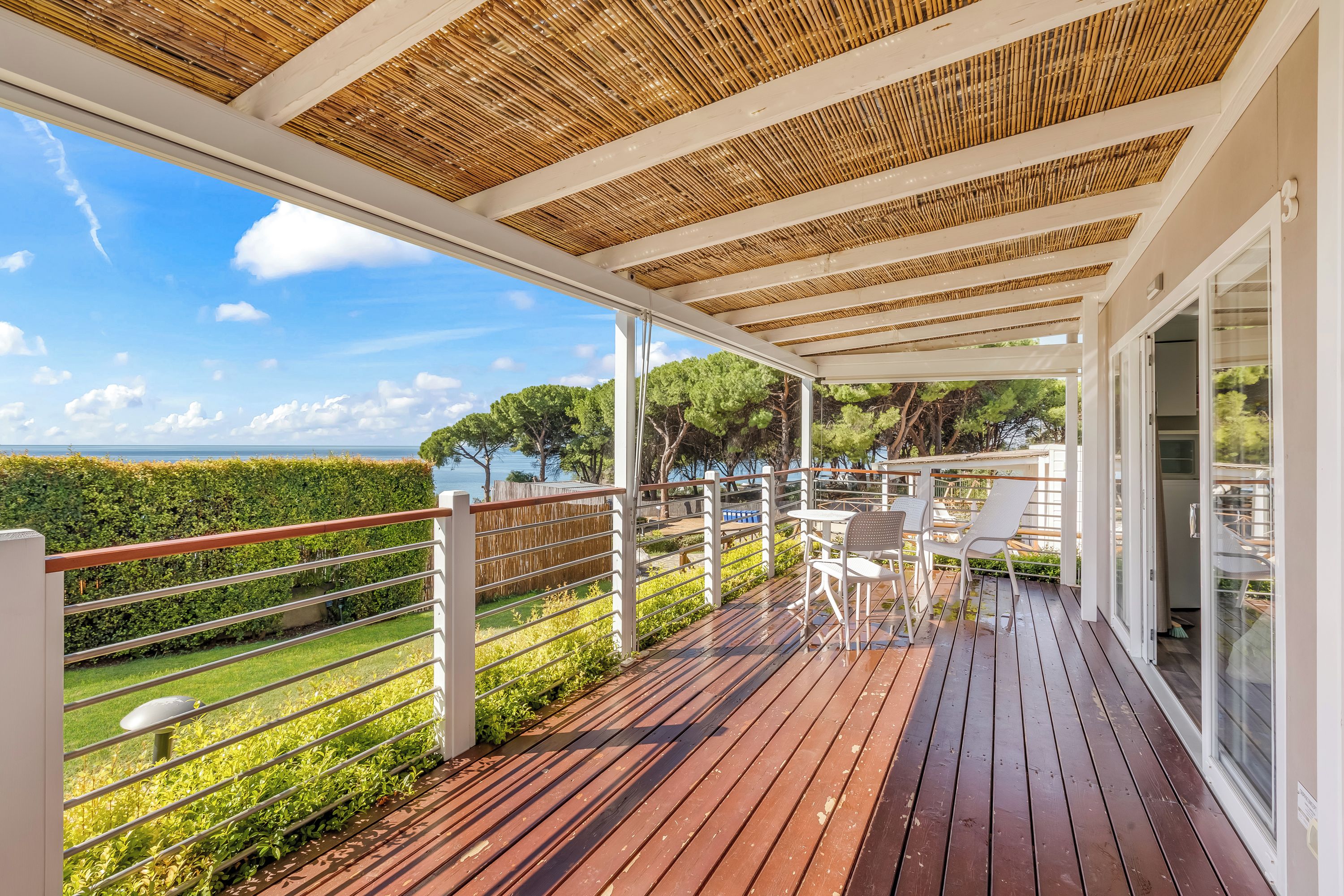 Une terrasse extérieure couverte avec vue sur la mer, un plancher en bois et des meubles d’extérieur blancs.