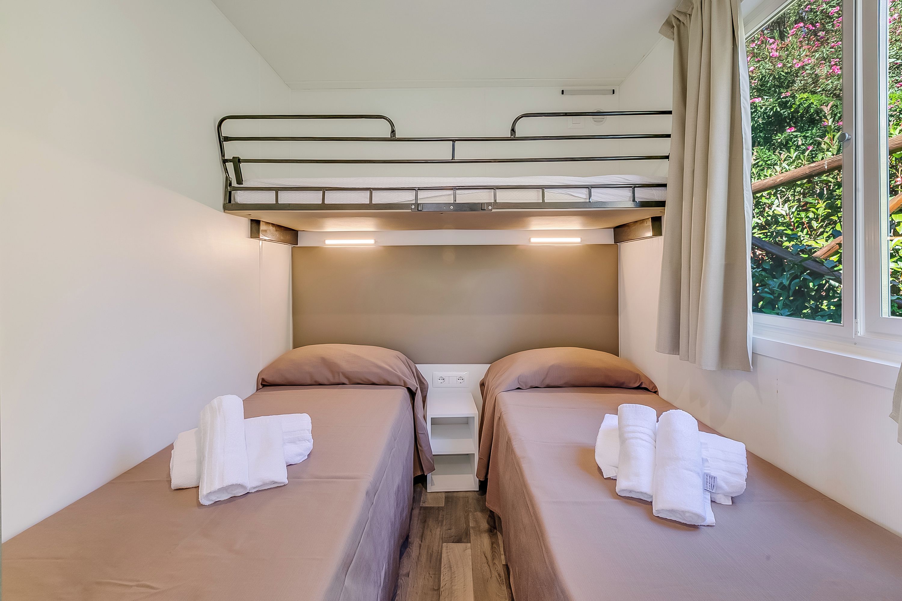 Une chambre avec un lit superposé, comportant deux lits, chacun avec un oreiller et une serviette enroulée. Il y a une fenêtre avec des rideaux et une vue sur la verdure à l’extérieur.