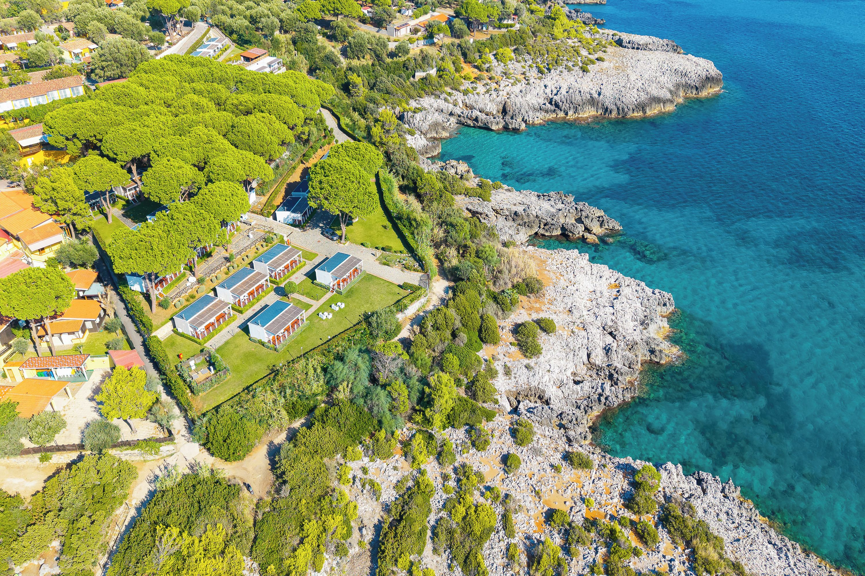 An aerial view of a coastal property with a rocky shoreline, a green park area, and several buildings with solar panels.
