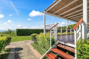 Un espace extérieur abrité, doté d’un escalier en bois menant à une terrasse, le tout entouré de verdure et d’un chemin en pierre.