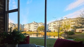 A room with a large window overlooking a lake, a potted plant, and a red chair. - Sweet Magnolias | Beach Escape Near Top Courses (Myrtle Beach)