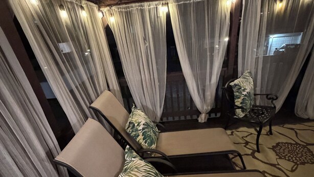 Un espacio de estar al aire libre techado con sillas acolchadas y una mesa, decorado con cortinas de gasa y guirnaldas de luces.