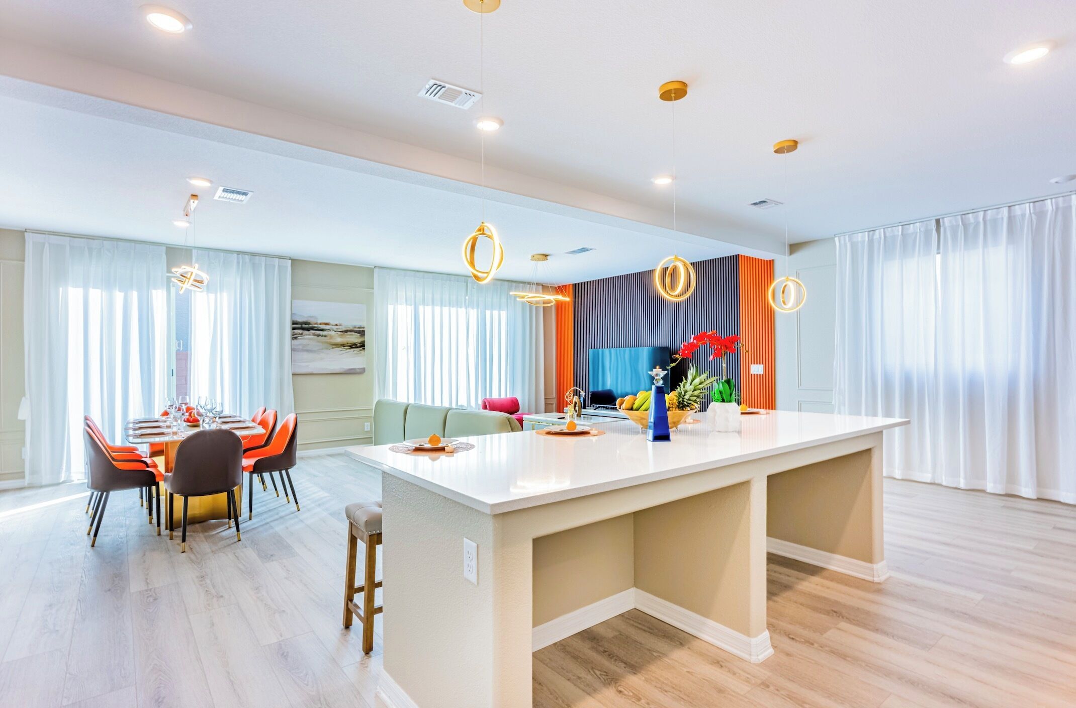 Une cuisine moderne avec un îlot central, un coin repas avec des chaises orange et un salon avec un canapé et un téléviseur.
