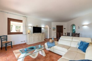 A living room with a sofa, a glass coffee table, a television on a cabinet, and a dining area with chairs and a table. - Villa White & Blue - Private Pool (Campanedda)