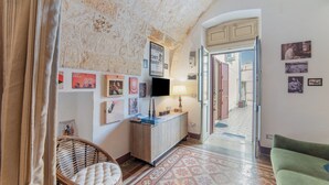 Une pièce avec un plafond en pierre, un canapé vert, un fauteuil en osier, une armoire en bois et un sol à motifs.
