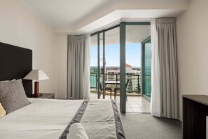 Een hotelkamer met een bed, een nachtkastje, een lamp, een balkon met een tafel en stoelen, en uitzicht op de stad.
