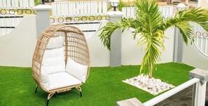 Una silla de mimbre con cojines blancos sobre césped artificial, una palmera y una cerca blanca con un portón.