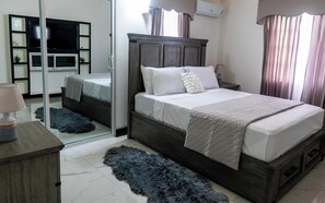 Un dormitorio con cama, mesita de noche, espejo, armario y ventana con cortinas.