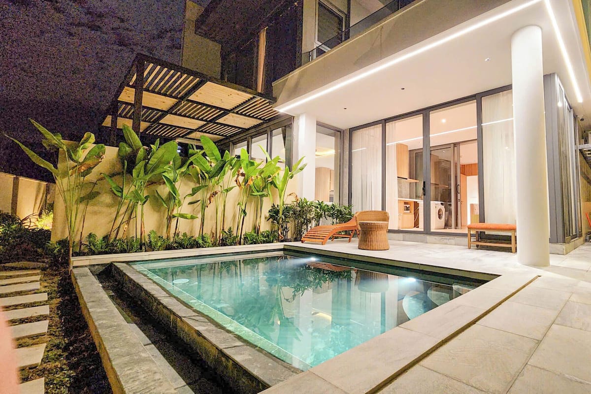 Un área de piscina moderna al aire libre con cerramiento de vidrio, rodeada de plantas tropicales y un patio con mobiliario.