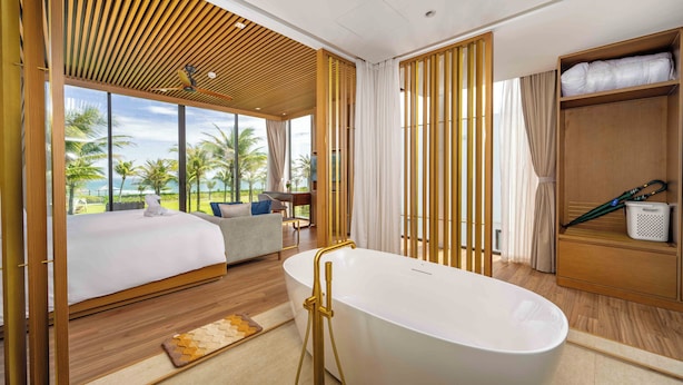 A modern hotel room with a large bed, a bathtub, a sofa, and a view of palm trees. - Regal Riverside Villas (Da Nang)
