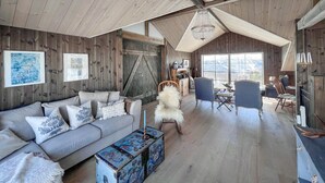 Ein gemütliches Blockhaus-Interieur mit Holzbalkendecke, einer Couch mit gemusterten Kissen, einem weißen, flauschigen Sessel und einem Tisch mit einer dekorativen Schatulle.