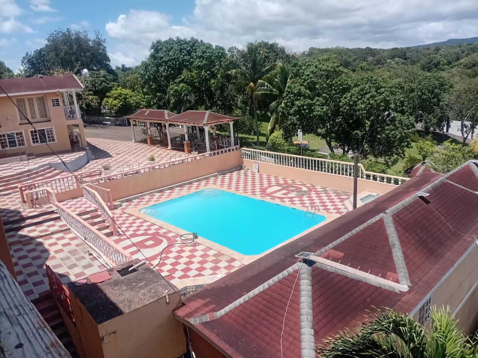 A swimming pool with a checkered pattern around it, a covered seating area, and a view of surrounding greenery.
