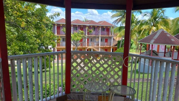 Un balcon couvert, agrémenté d’une balustrade en treillis, d’une table et de chaises, qui offre une vue sur un immeuble doté de nombreux balcons et bordé de palmiers.