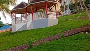 Un kiosque doté d’un toit rouge et de panneaux en treillis blancs, entouré d’une pelouse bien entretenue et de palmiers.