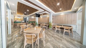 A modern dining area with wooden tables and chairs, a flat-screen TV mounted on the wall, and a kitchen in the background. - Horison IJ Kudus (Kudus)