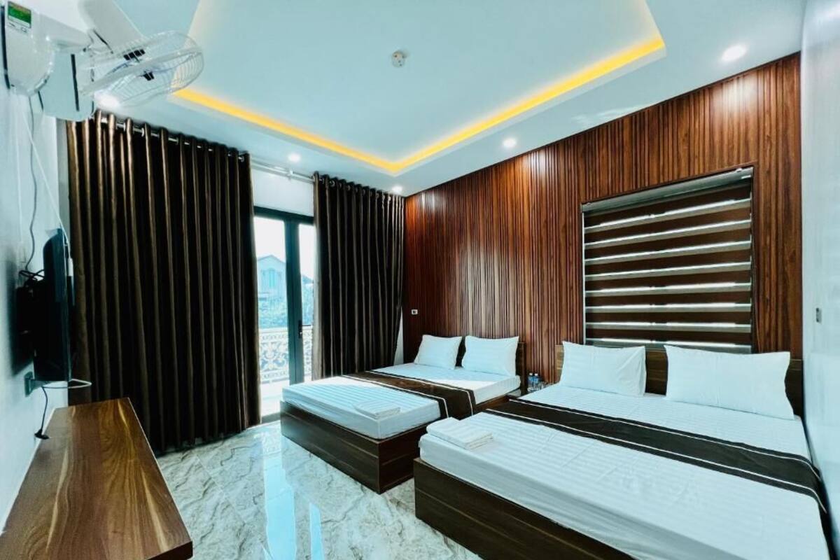 A hotel room with two beds, a wooden desk, a large window with curtains, and a ceiling fan.
