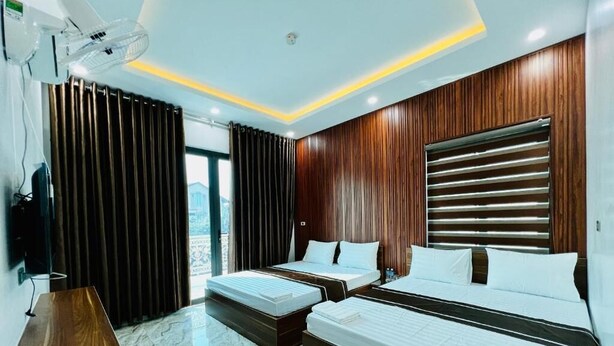 Une chambre d’hôtel avec deux lits, un bureau en bois, une grande fenêtre avec des rideaux et un ventilateur de plafond.