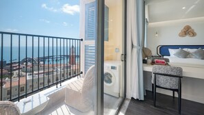 Een balkon met uitzicht op zee en gebouwen, een wasmachine en een bed met hoofdbord en kussens.