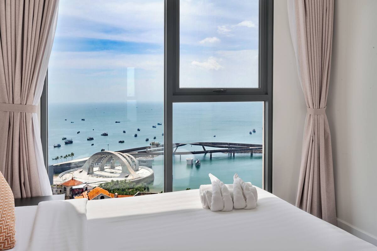 Ein Hotelzimmer mit großem Fenster und Blick auf das Meer, Boote und eine Brücke.
