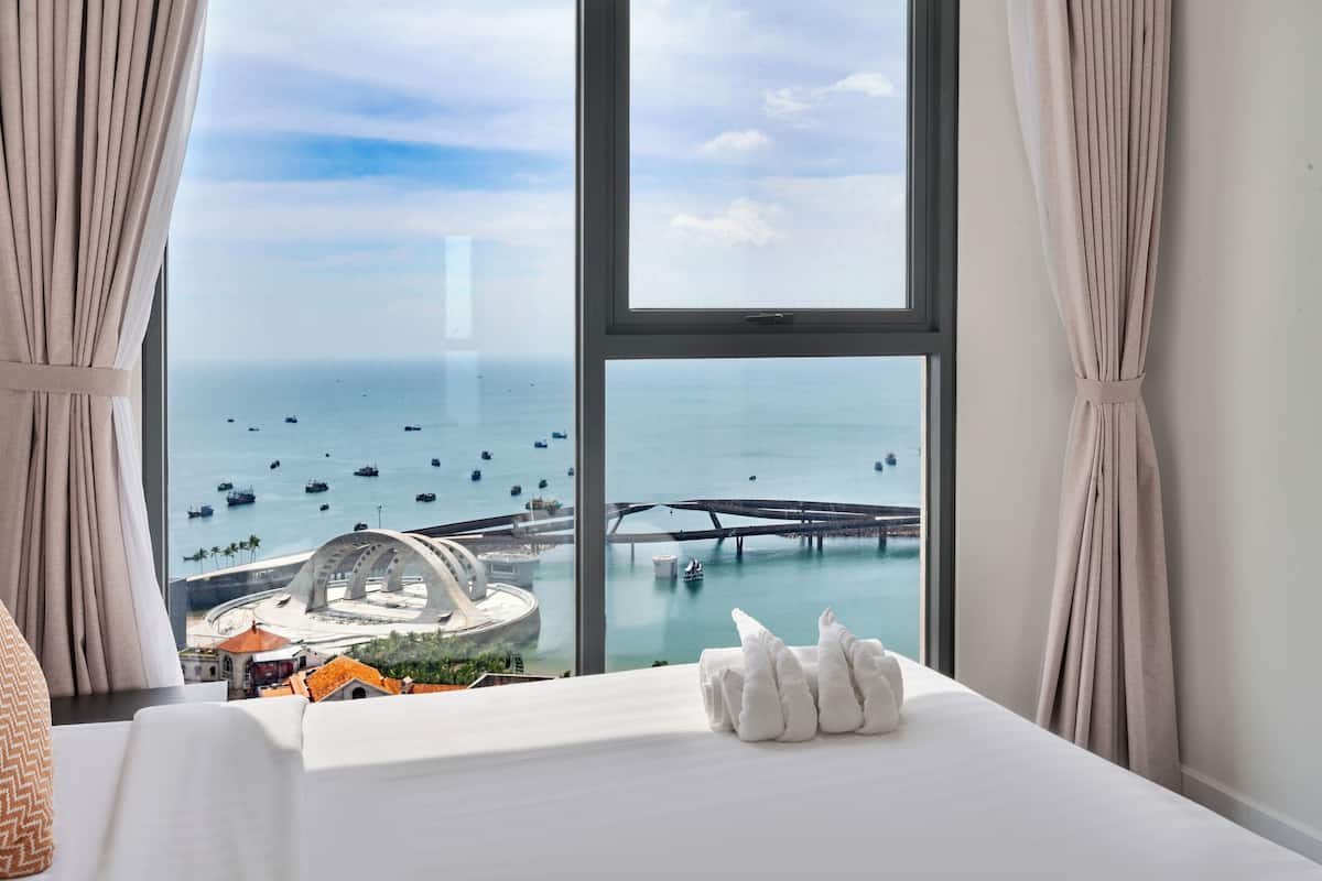 A hotel room with a large window overlooking a marina and a bridge, a neatly made bed, and a view of a distinctive building structure.