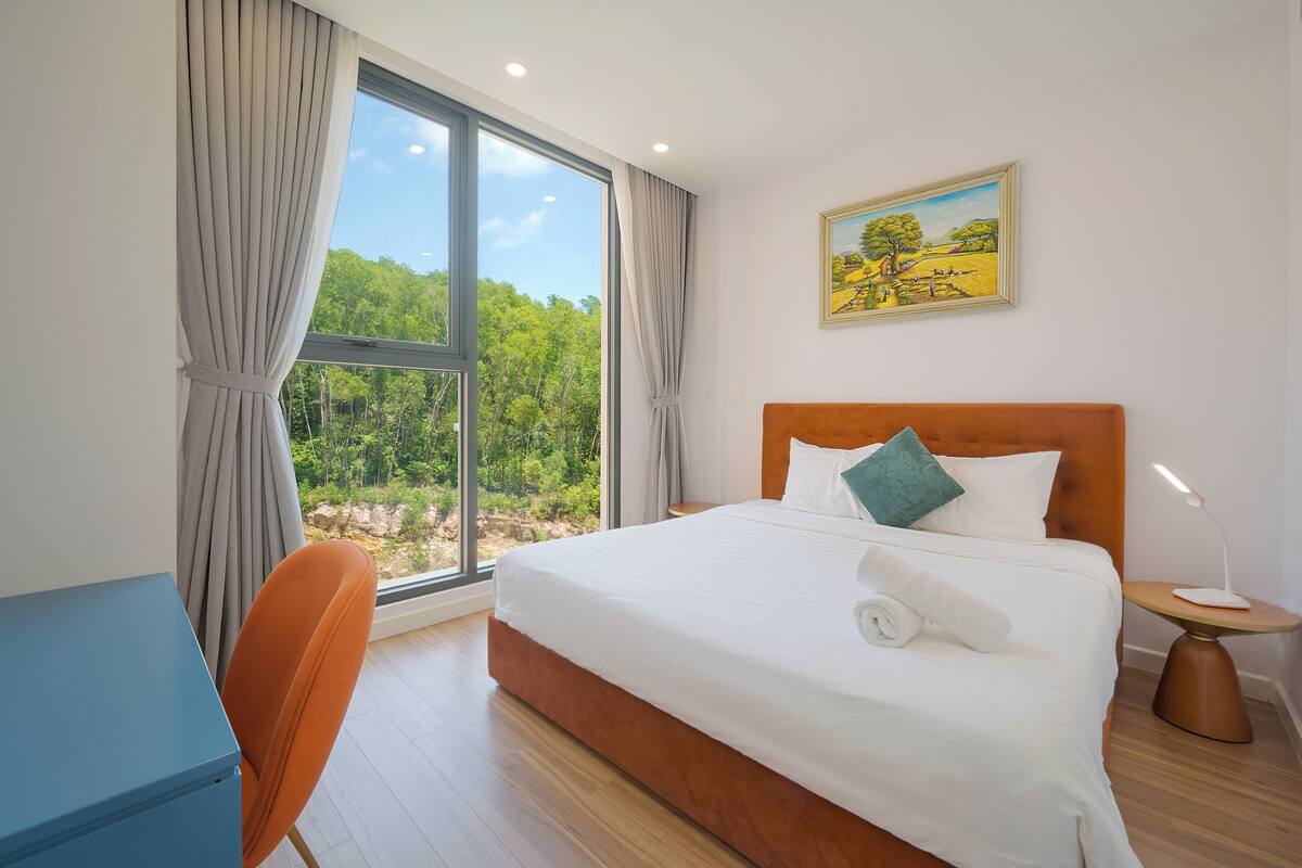 Ein Hotelzimmer mit großem Fenster, einem Bett mit weißer Bettwäsche, einem Holzkopfstück, einem Schreibtisch und einem Bild an der Wand.