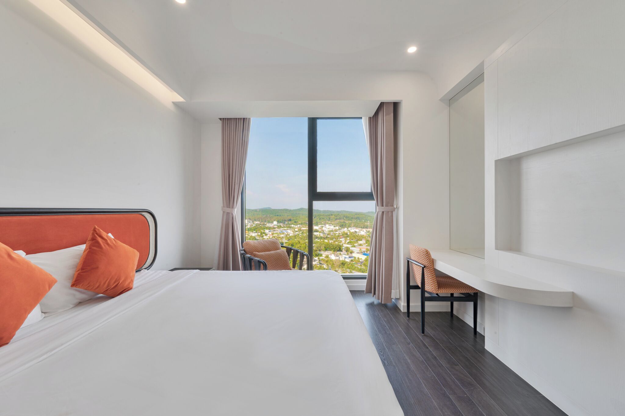 A hotel room with a large bed, a desk, and a view of a cityscape through a large window.