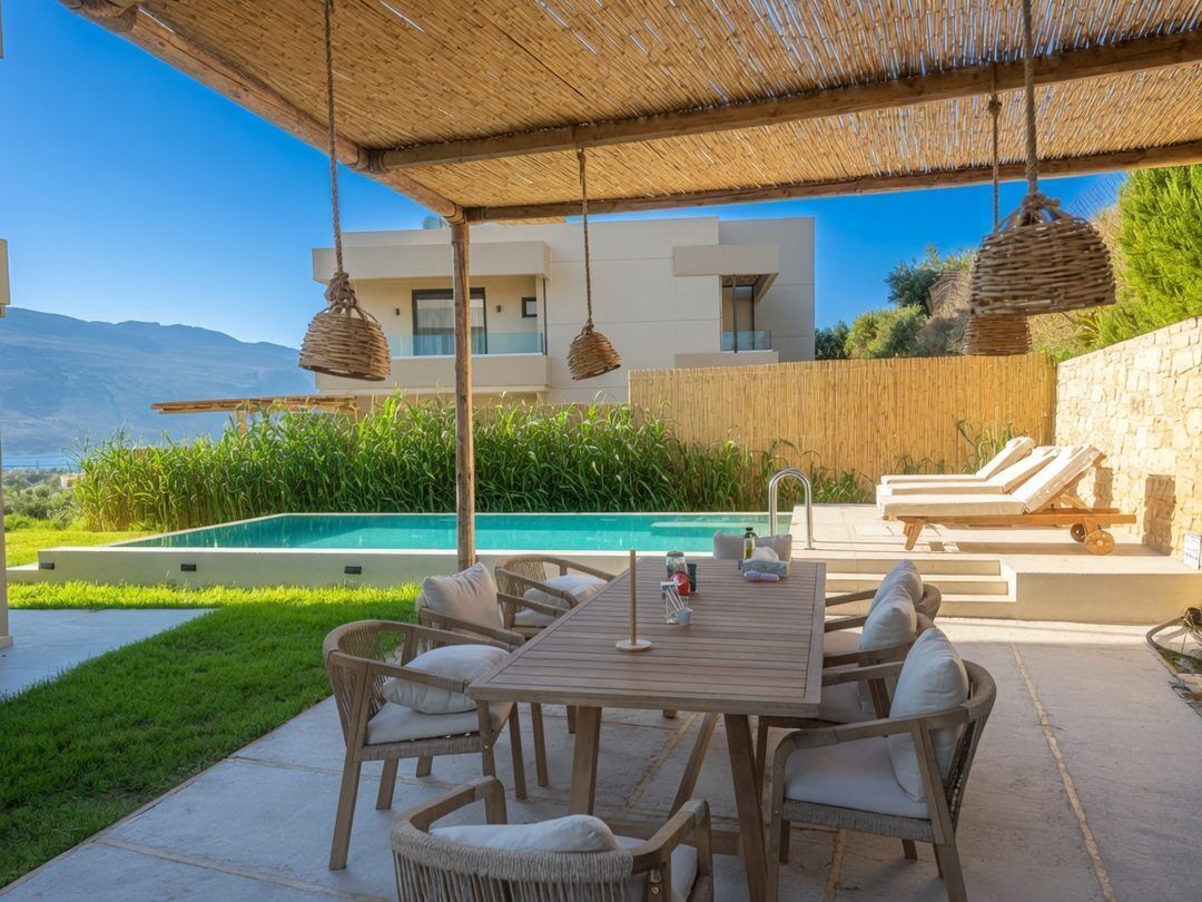 A poolside area with a wooden table and wicker chairs, a stone wall, and a view of a mountain.