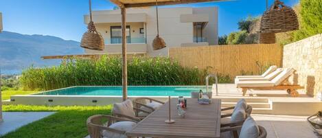 A poolside area with a wooden table and wicker chairs, a stone wall, and a view of a mountain.
