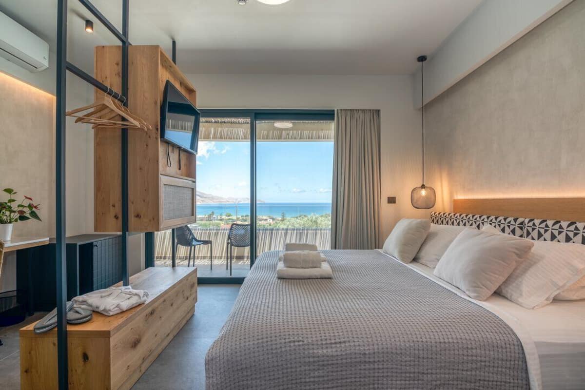 A bedroom with a large bed, a wooden bench, a TV, and a balcony with a view.
