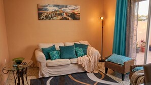 Ein Wohnzimmer mit einer Couch, einem Korbsessel, einem Glastisch, einer Stehlampe und einem Gemälde an der Wand.