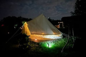 A tent with a conical roof structure, supported by poles, and illuminated by internal lighting.