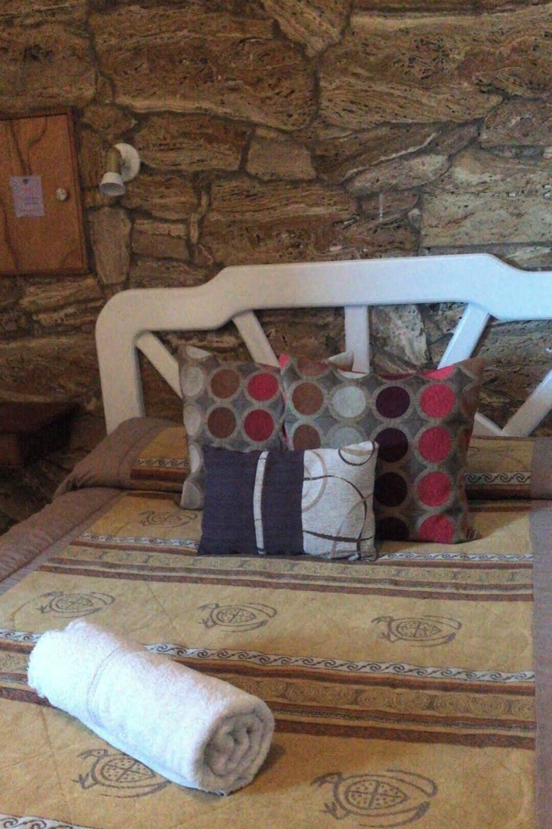 A bed with a patterned bedspread, a rolled towel, and decorative pillows against a stone wall.