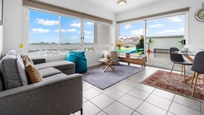 Ein modernes Wohnzimmer mit Sofa, Couchtisch und Fernseher, große Fenster mit Blick auf das Wasser und ein Balkon mit Essbereich.