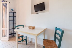 Ein Zimmer mit einem weißen Tisch, zwei türkisfarbenen Stühlen, einem Korbwannenmöbel und einem Wand-Fernseher sowie einem Eckregal.