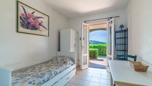 Una habitació amb un llit, una taula, una cadira, un armari i una porta que dona a un jardí.