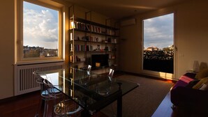 Un salon moderne avec une table en verre, des chaises transparentes, une bibliothèque, un téléviseur et une grande fenêtre donnant sur la ville.