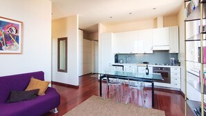 Un appartement moderne avec un canapé violet, une table à manger en verre, des meubles de cuisine blancs et une bibliothèque.