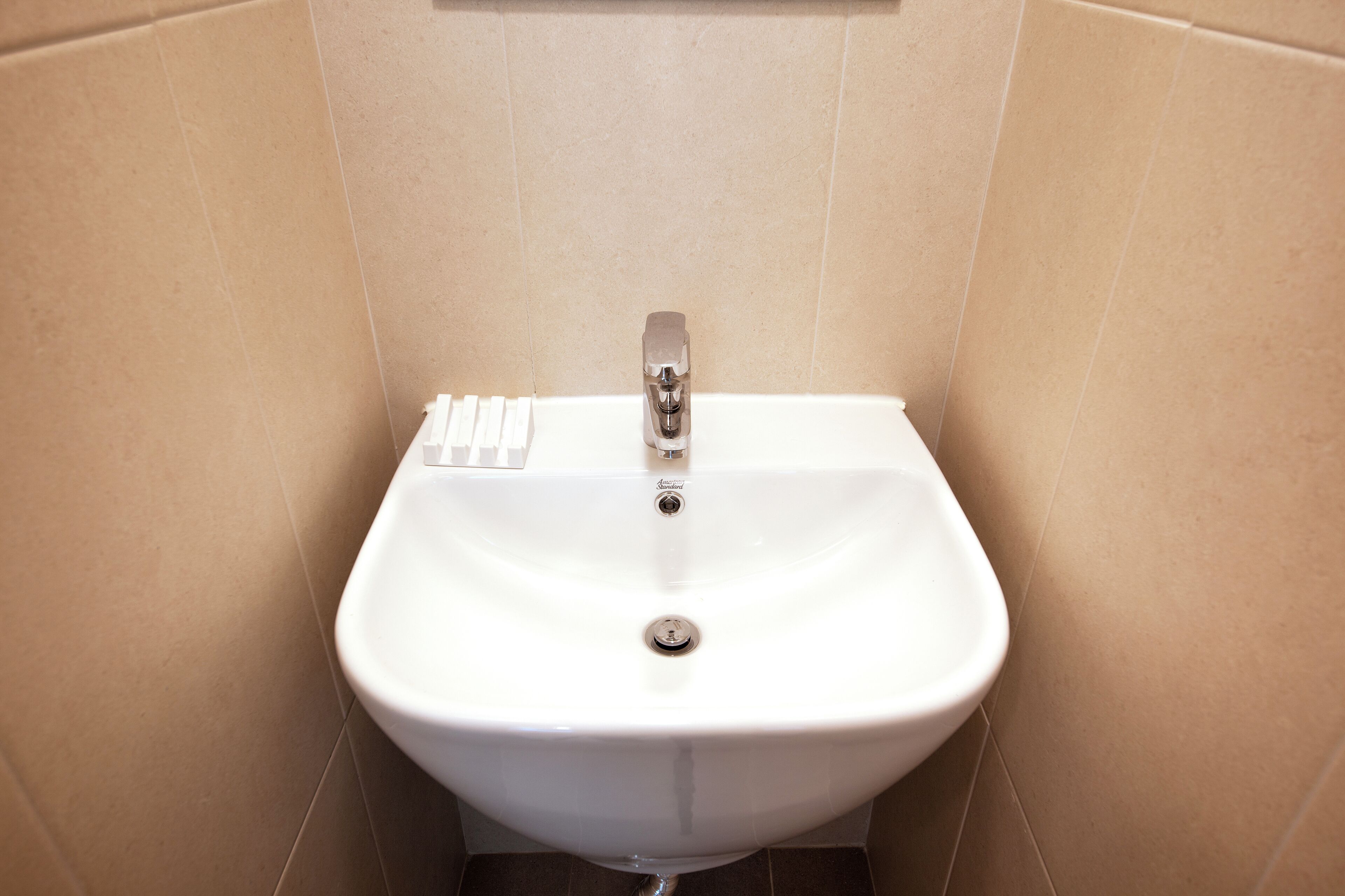 Un lavamanos pedestal blanco con un solo grifo y tapón, frente a paredes de azulejos color beige.