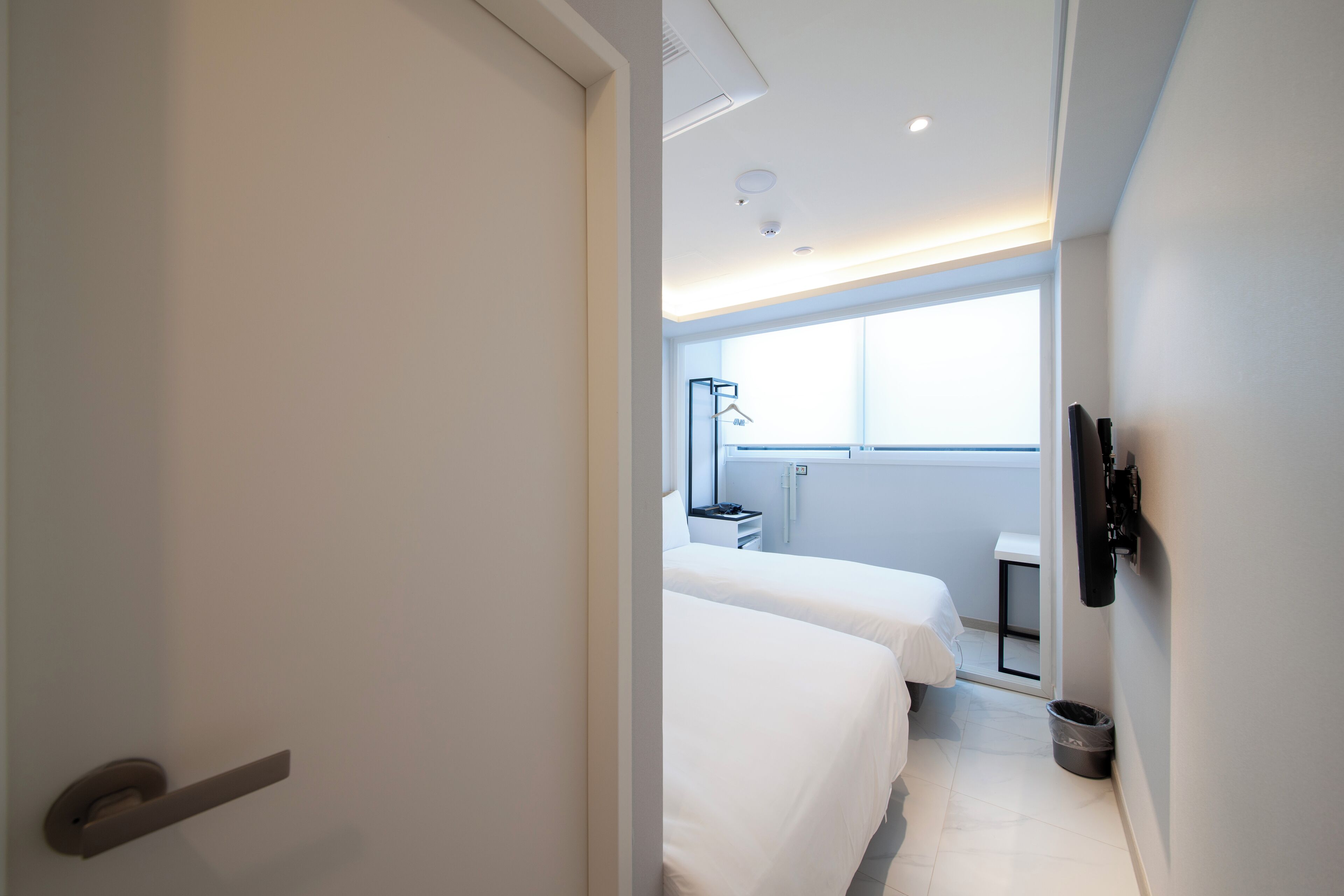 Camera d'albergo con due letti, un comodino, una piccola scrivania, una sedia, una finestra e una porta.