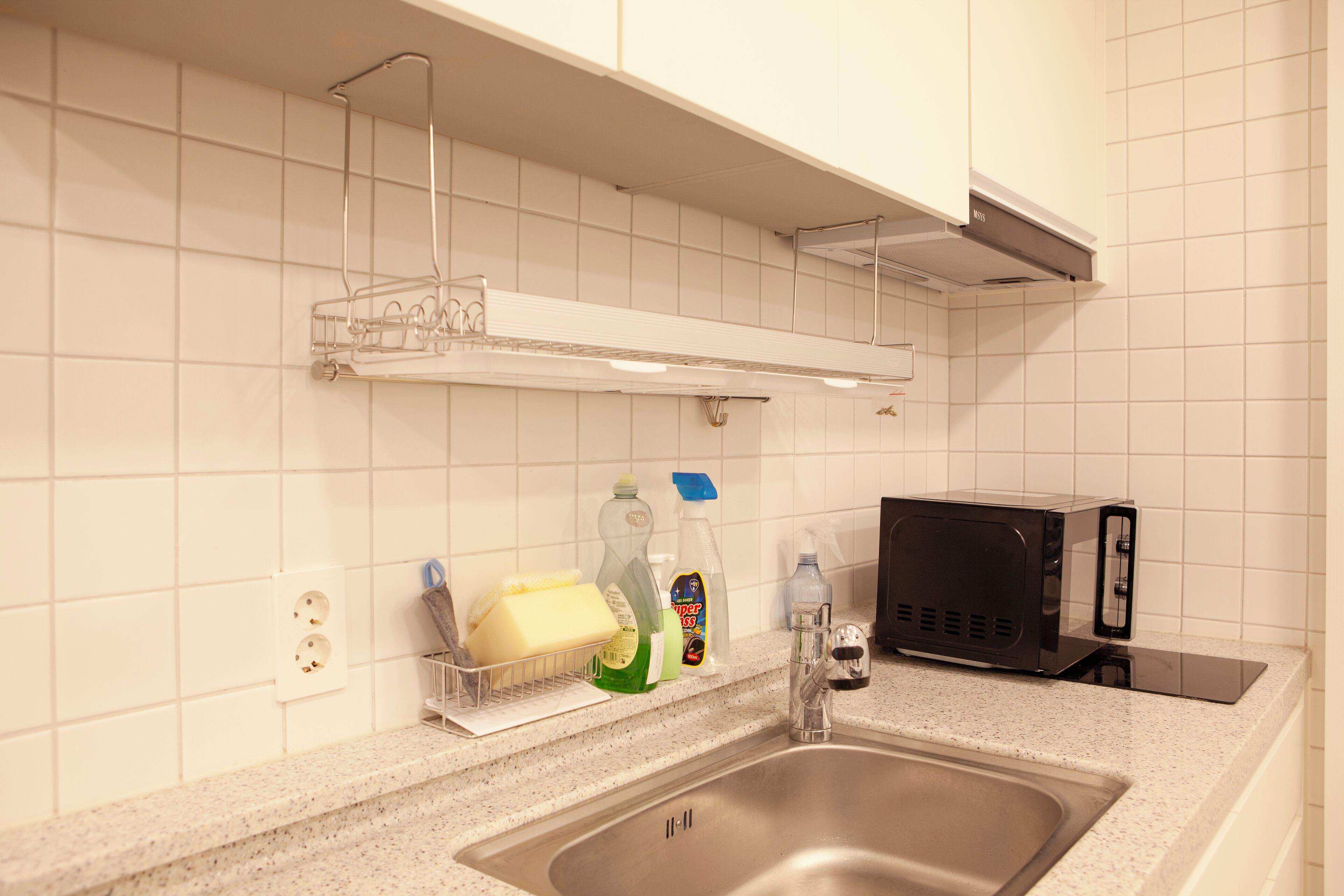 Una cocina con un lavamanos de acero inoxidable, un microondas y un estante con productos de limpieza.