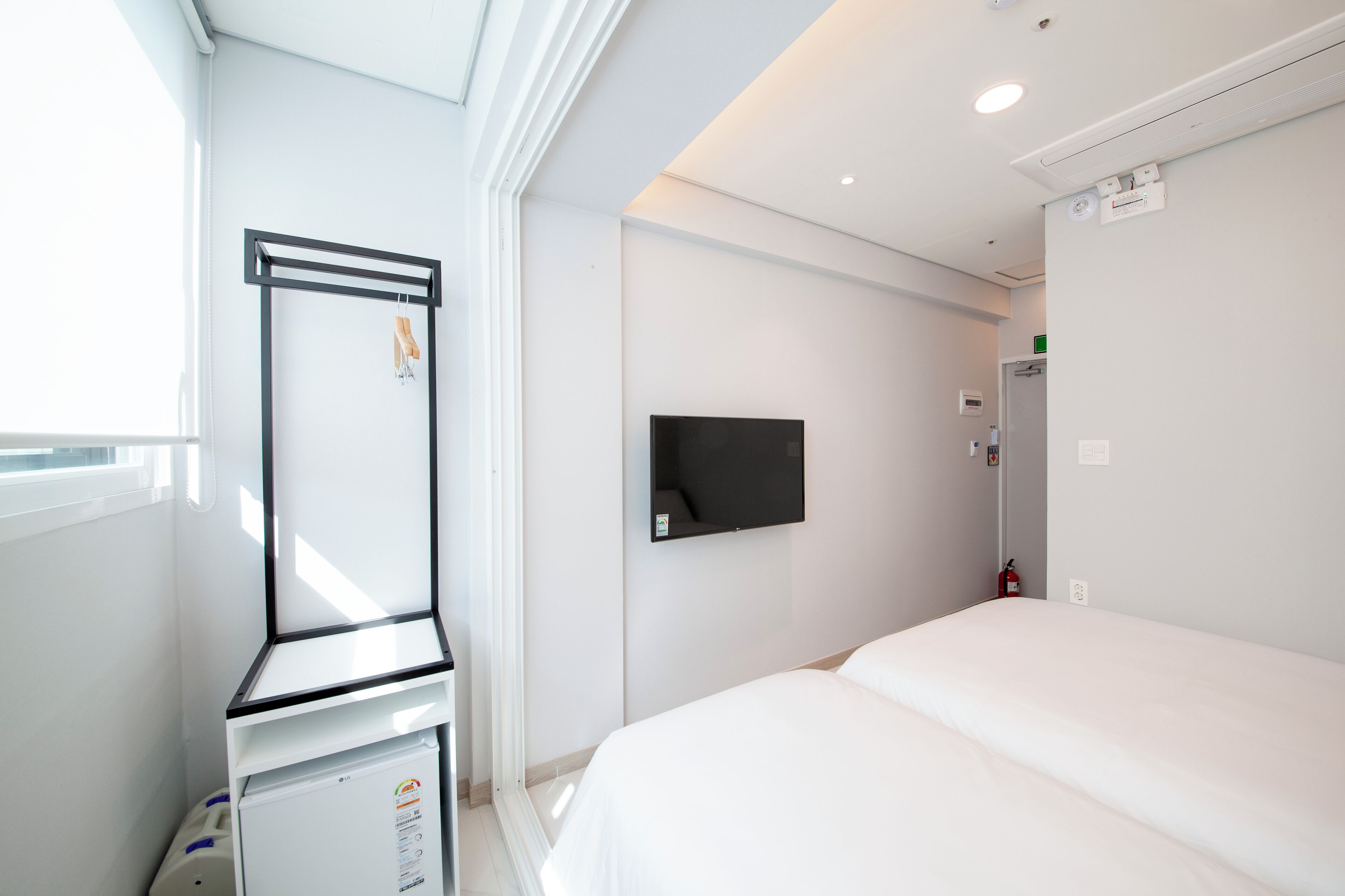 A hotel room with a bed, a flat-screen TV mounted on the wall, a small refrigerator, and a window with blinds.