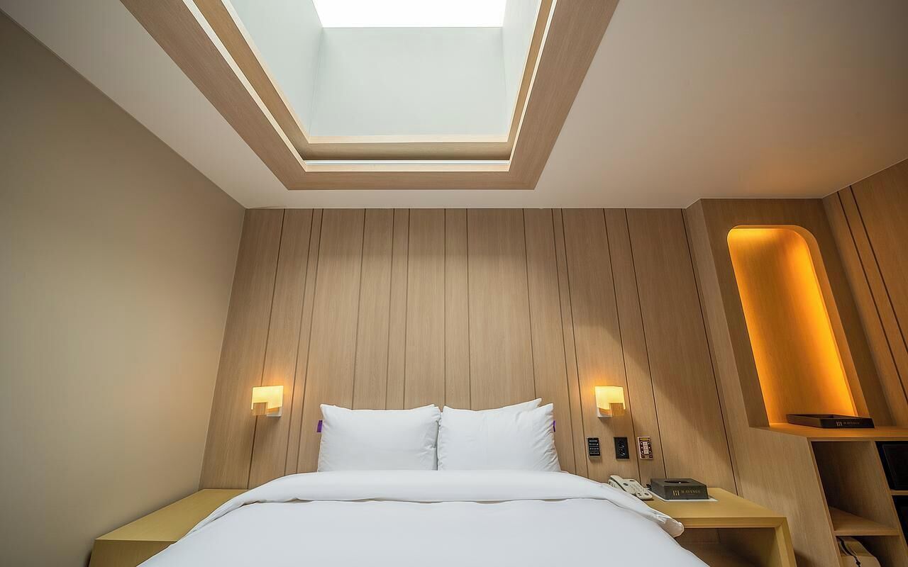 A modern hotel room with a skylight, wooden panel walls, a bed with white linens, bedside lamps, and a nightstand with a phone and a remote control.