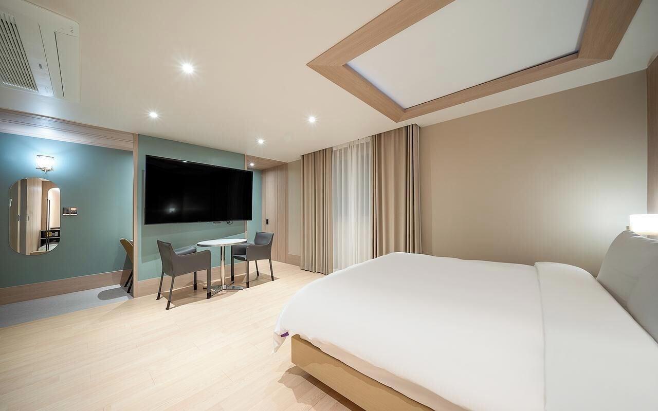 Habitación de hotel con cama, escritorio con silla, televisor y ventana grande con cortinas.