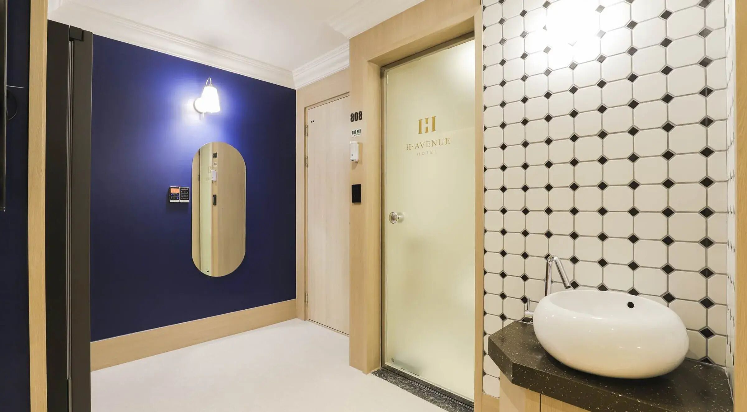 A hotel room with a blue wall, a door labeled 'H. Avenue Hotel,' and a bathroom with a white sink and hexagonal tile backsplash.