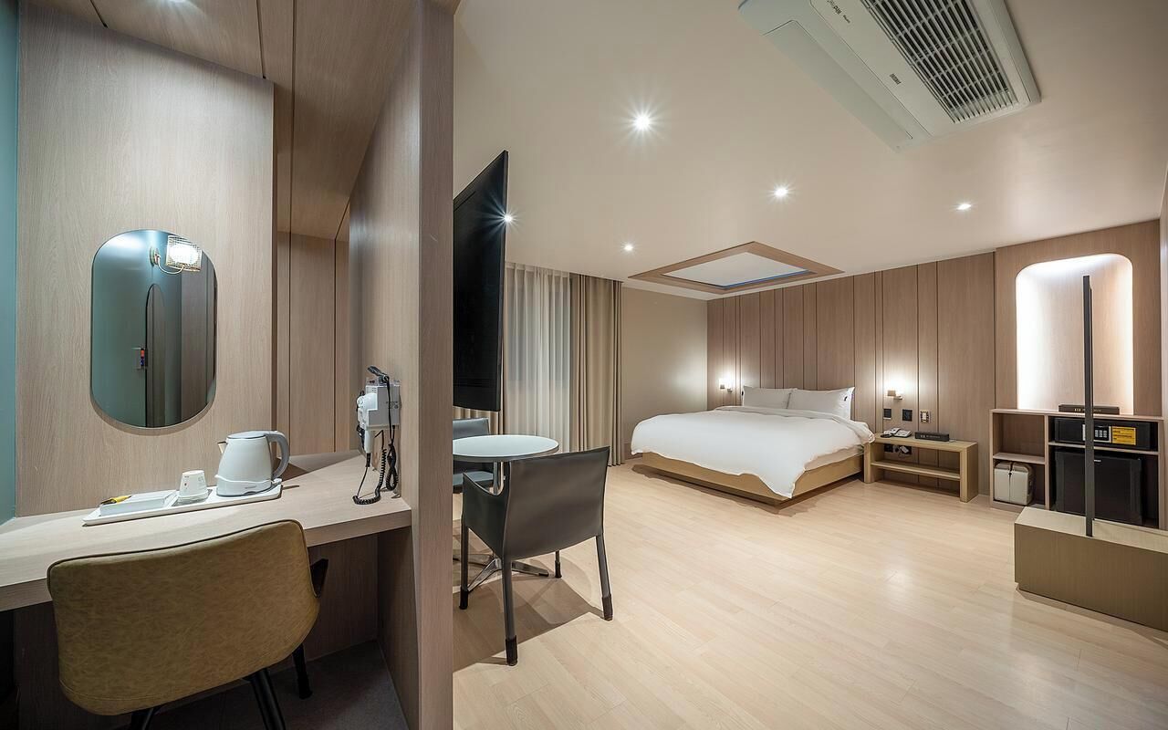 A modern hotel room with a desk, chair, bed, bedside table, and a TV.