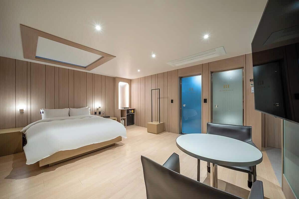 Una habitación de hotel moderna con cama, mesitas de noche, escritorio y silla. Hay dos puertas, una con panel de vidrio y otra con panel de vidrio esmerilado.