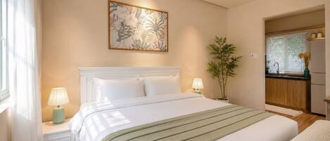 A hotel room with a bed, bedside lamps, a wall-mounted artwork, a potted plant, and a view of the kitchen.