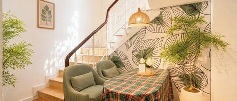 Una escalera con barandilla de madera, un sillón verde, una mesa cubierta con mantel escocés y una planta en maceta.