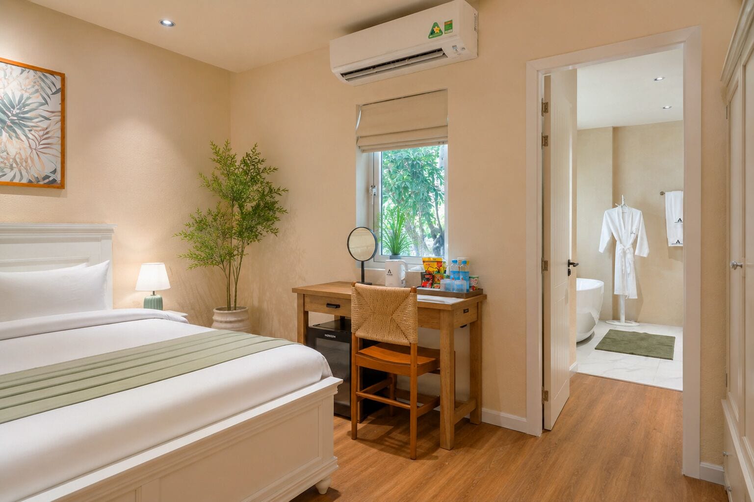 A hotel room with a bed, a desk with a chair, a potted plant, a window with a view of greenery, and an air conditioning unit.