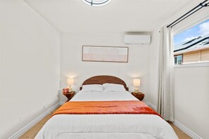 A bedroom with a wooden bed, white walls, a ceiling light, a window with curtains, and a wall-mounted air conditioner. - 2-bedroom house in nice Tustin with cool AC for relaxing stays (Tustin)